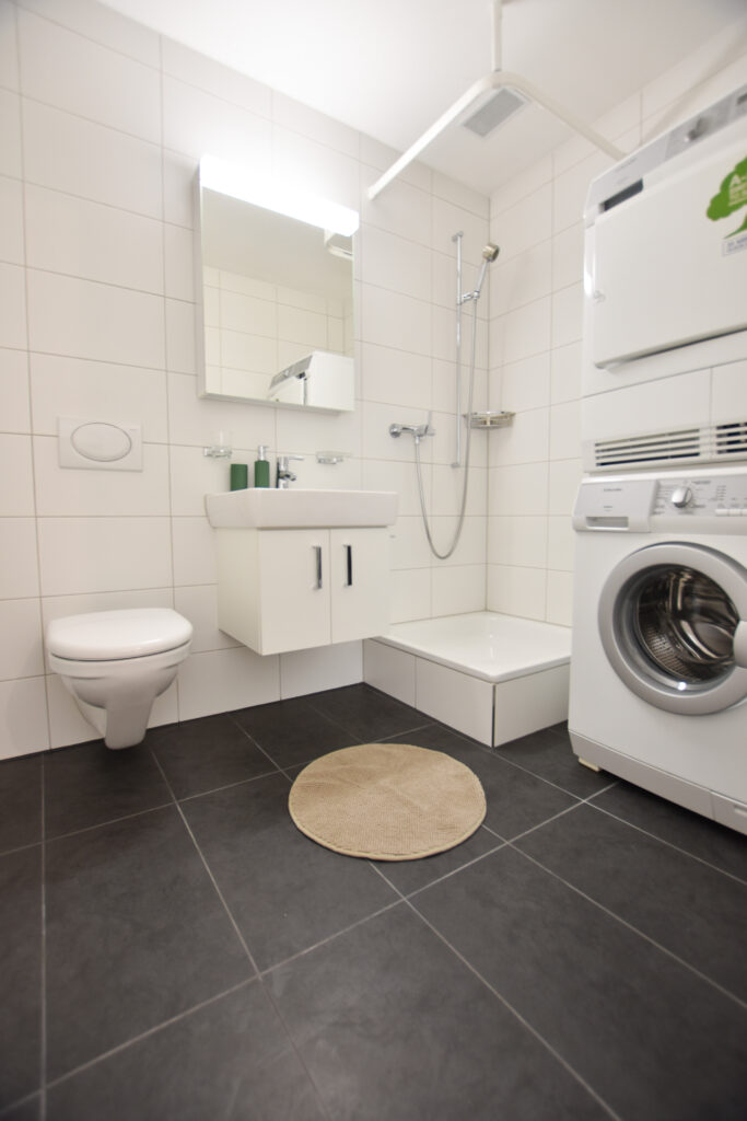 Une salle de bain moderne avec des carreaux blancs. Elle dispose d'un WC suspendu, d'un petit lavabo surmonté d'un miroir et d'un espace douche. À côté de la douche se trouve une unité de machine à laver et de sèche-linge empilée l'une sur l'autre. Un tapis rond beige est posé sur le sol carrelé foncé.