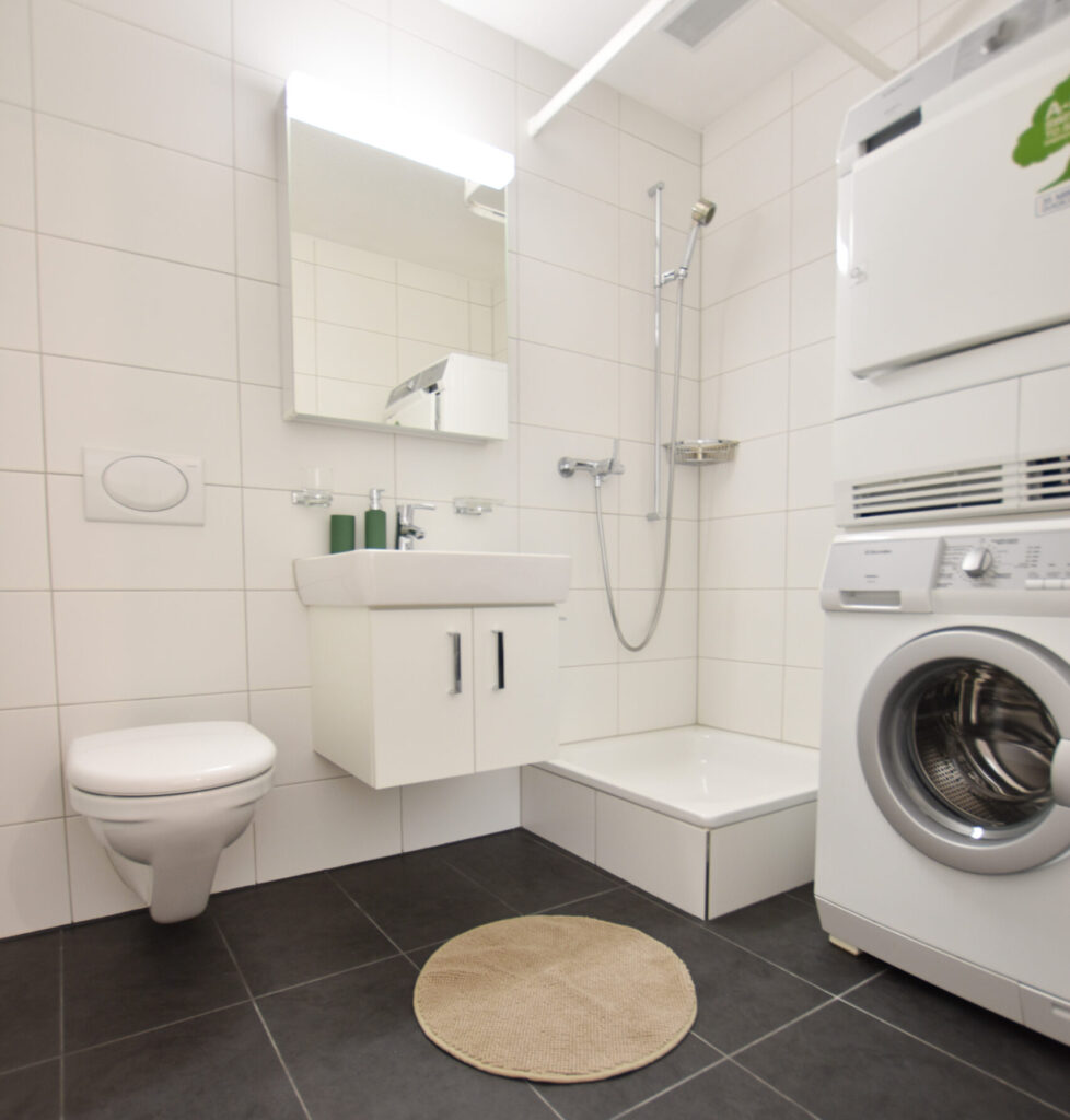 Ein modernes Badezimmer mit weißen Fliesen. Es verfügt über eine Wandtoilette, ein kleines Waschbecken mit einem Spiegel darüber und einen Duschbereich. Neben der Dusche steht eine übereinander gestapelte Waschmaschinen- und Trocknereinheit. Auf dem dunklen Fliesenboden liegt eine runde beige Matte.