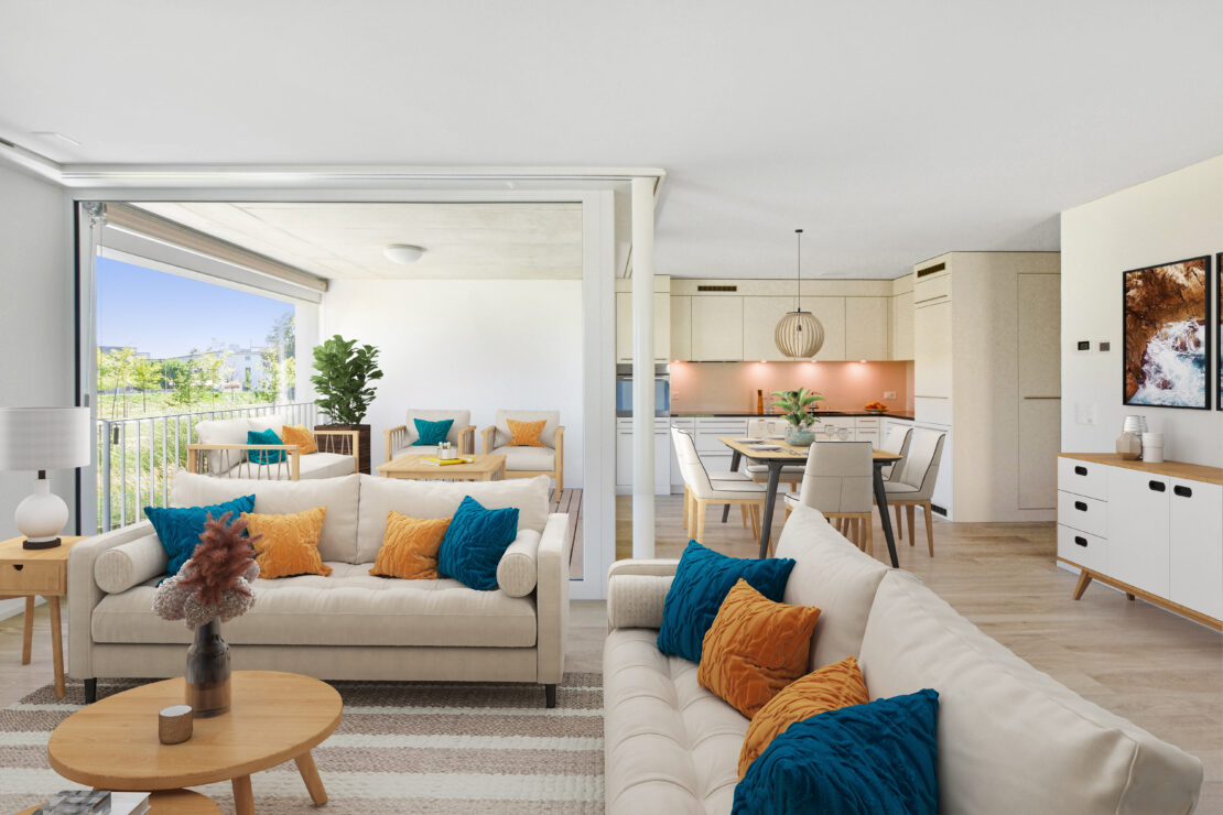 Ein moderner, offener Wohn- und Essbereich mit beigen Sofas, bunten Kissen und einem Couchtisch aus Holz. Der Essbereich verfügt über einen Holztisch und Stühle. Ein Balkon mit Sitzgelegenheiten im Freien ist sichtbar und bietet einen Blick ins Grüne.