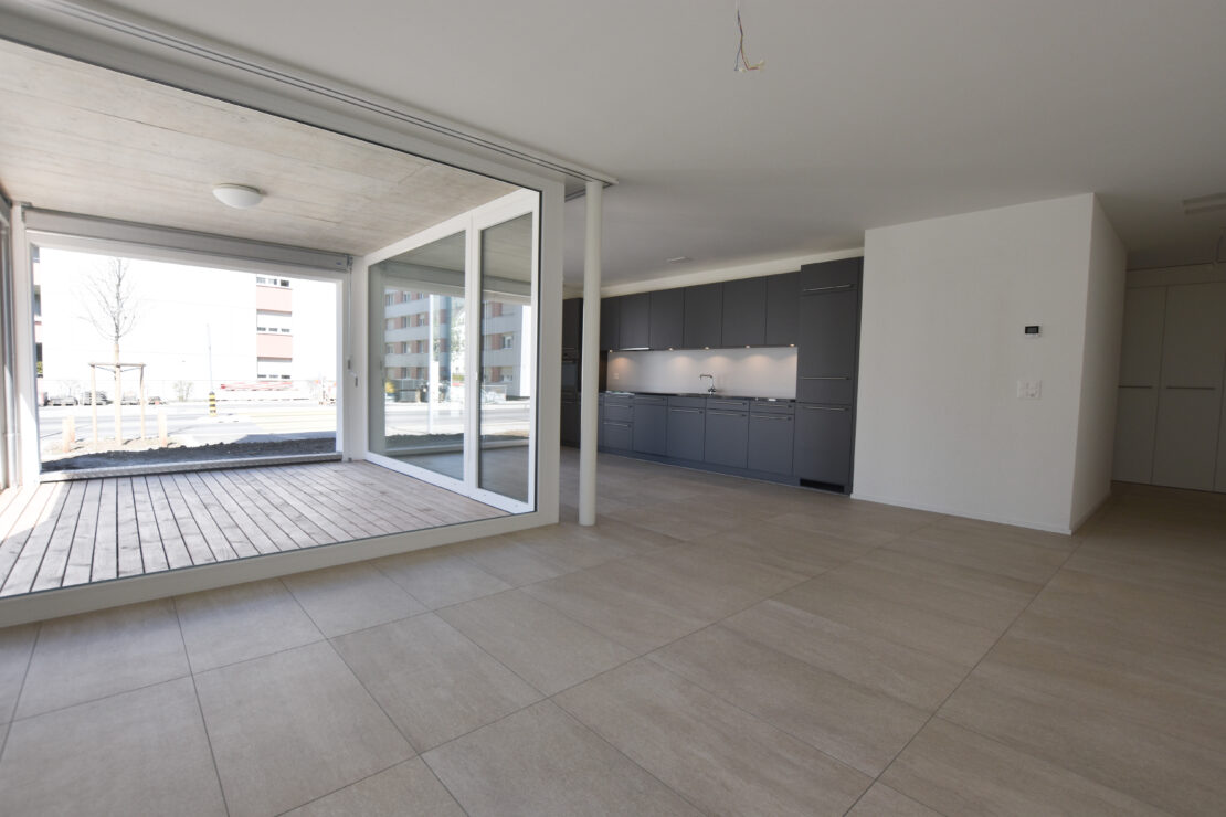Eine moderne, leere Wohnung mit hellbeigem Fliesenboden, grosszügigem, offenem Grundriss und grossen Fenstern mit Blick auf eine kleine Terrasse mit Holzbelag. Der Küchenbereich ist mit eleganten dunklen Schränken und Einbaugeräten ausgestattet.