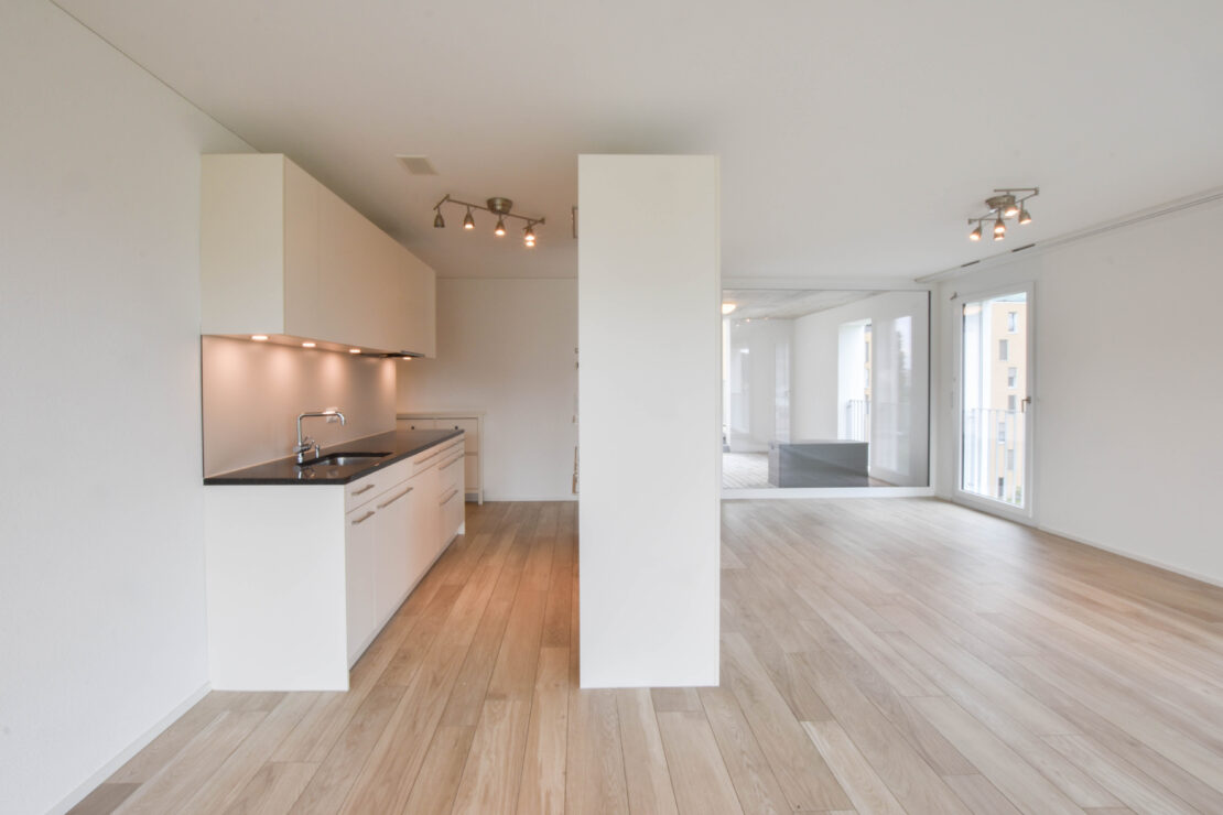 Eine geräumige, moderne Wohnung mit hellem Holzboden und weissen Wänden. Die Küche ist auf der linken Seite und verfügt über elegante Schränke und eine Arbeitsplatte mit eingebautem Herd. Grosse Fenster auf der rechten Seite lassen natürliches Licht herein und überall sind Deckenleuchten verteilt.
