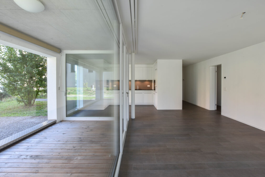 Eine moderne, leere Wohnung mit Braunem Fliesenboden, grosszügigem, offenem Grundriss und grossen Fenstern mit Blick auf eine kleine Terrasse mit Holzbelag. Der Küchenbereich ist mit eleganten weissen Schränken und Einbaugeräten ausgestattet.