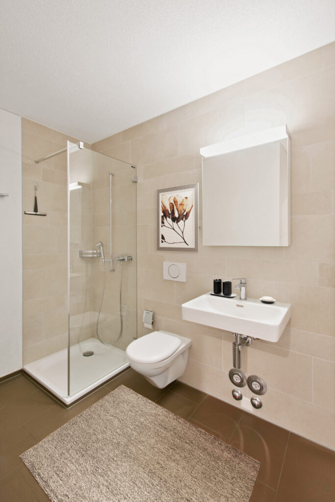 Une salle de bain moderne avec des carreaux beiges dispose d'une douche vitrée, de toilettes murales et d'un lavabo rectangulaire blanc. Une armoire à glace est suspendue au-dessus du lavabo et une œuvre d'art végétale décorative est accrochée au mur. Le sol marron est recouvert d'un tapis structuré.