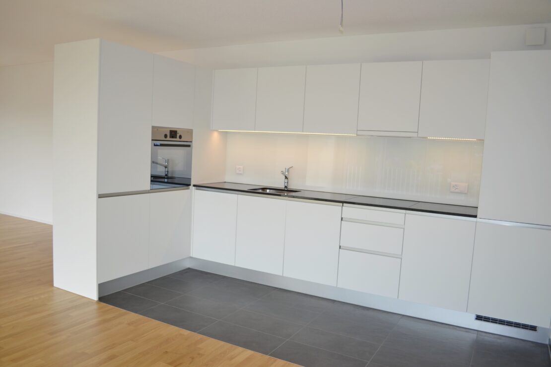 Eine moderne Küche mit eleganten weissen Schränken und Einbaugeräten, darunter ein Backofen. Die Arbeitsplatte ist schwarz und unter den Oberschränken verläuft eine LED-Lichtleiste. Der Boden besteht aus einer Kombination aus Holz und grauen Fliesen.