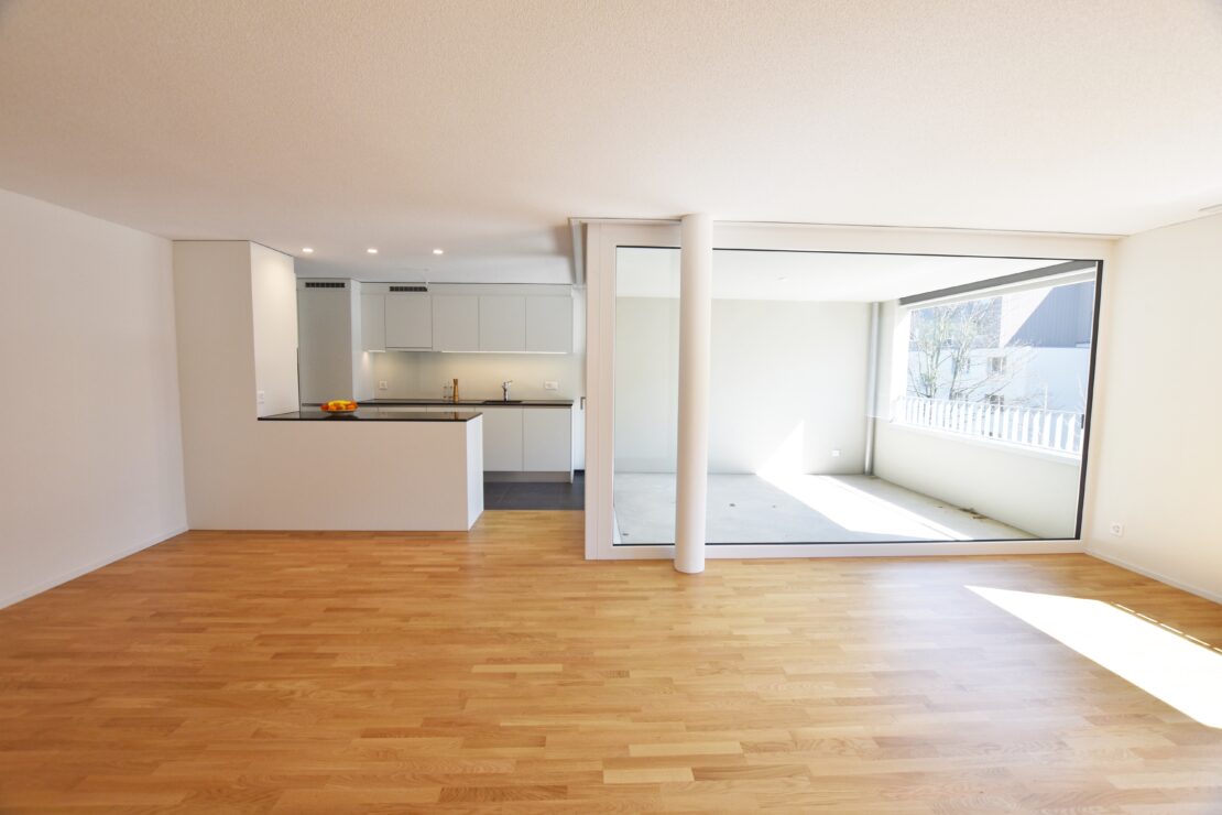Eine helle, moderne Wohnung verfügt über einen offenen Wohnbereich mit hellem Holzboden. Im Hintergrund ist eine elegante Küche mit weissen Schränken und dunkler Arbeitsplatte zu sehen. Ein grosses Fenster lässt reichlich natürliches Licht herein.