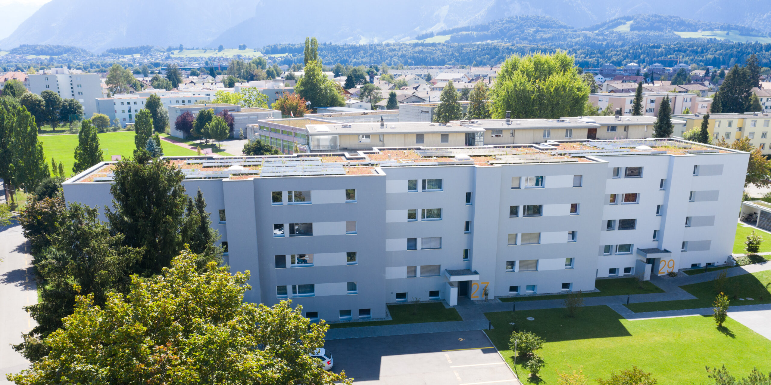 Luftaufnahme eines modernen Wohnhauses mit Flachdach und Sonnenkollektoren. Das Gebäude ist von grünen Rasenflächen, Bäumen und einem Parkplatz umgeben. Im Hintergrund sind in der Ferne Berge und ein klarer blauer Himmel zu sehen.