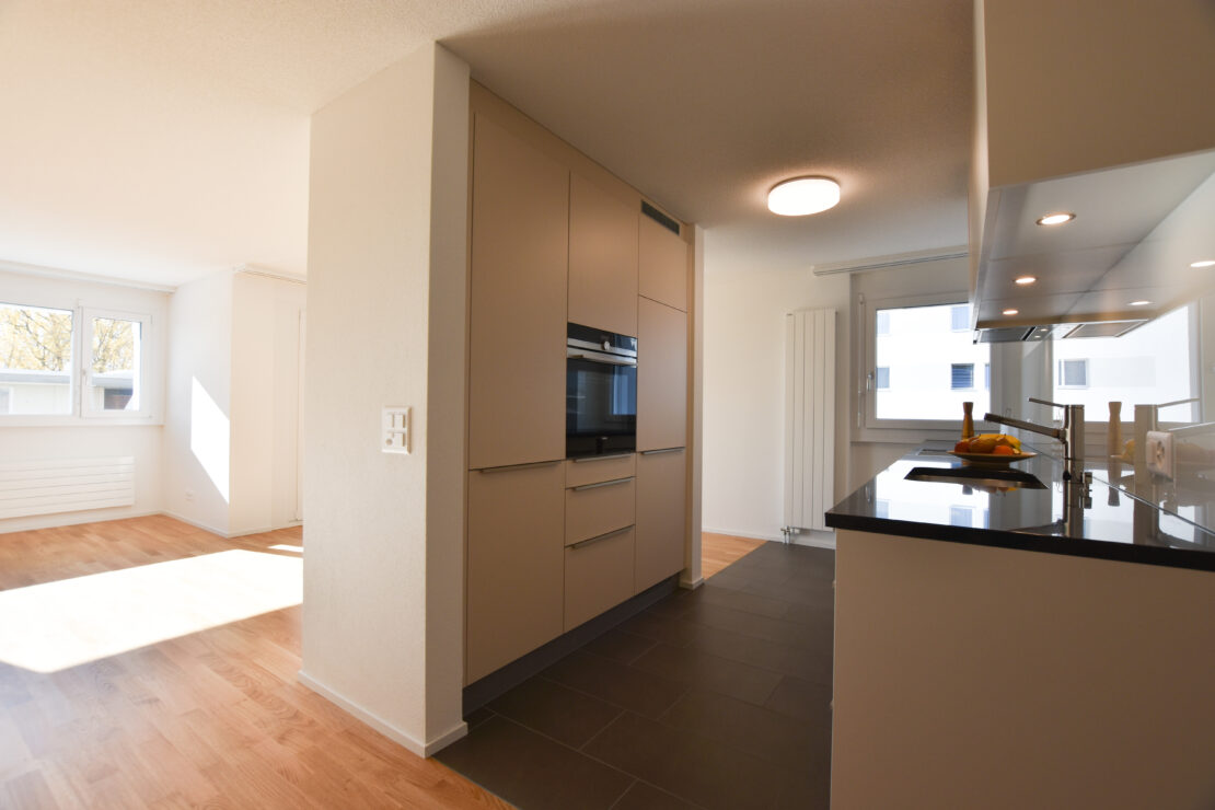 Eine moderne Küche mit beigen Schränken, einer schwarzen Arbeitsplatte und Einbaugeräten. Sonnenlicht strömt durch ein Fenster herein und erhellt den geräumigen angrenzenden Wohnbereich mit Holzboden.