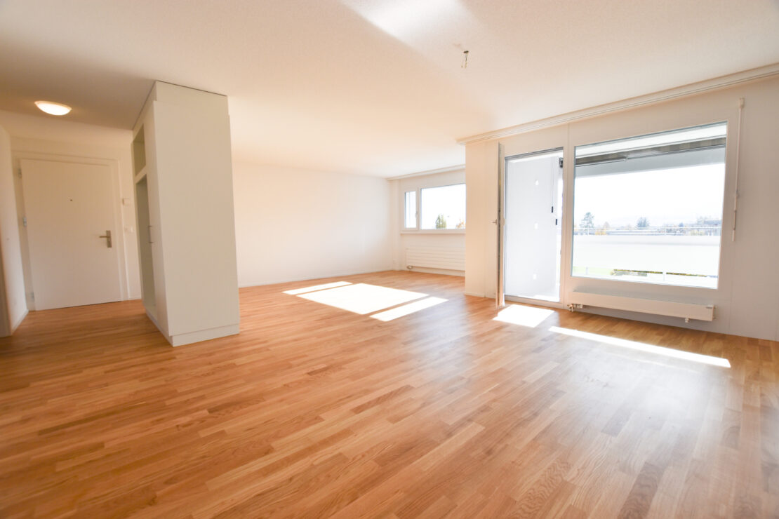 Heller, leerer Raum mit Hartholzboden, grossen Fenstern und einer Glastür, die auf einen Balkon führt. Weisse Wände und Decken sorgen für ein geräumiges, luftiges Gefühl. Natürliches Licht durchflutet den Bereich und hebt den Boden und den offenen Raum hervor.