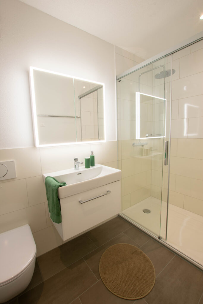 Salle de bain moderne avec douche vitrée, miroir éclairé et lavabo blanc. Une serviette verte est suspendue au-dessus du lavabo et un tapis rond beige est posé sur le sol carrelé gris. Des murs blancs complètent le design épuré et minimaliste.