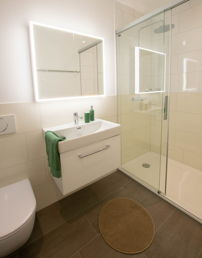 Modernes Badezimmer mit verglaster Dusche, beleuchtetem Spiegel und weissem Waschbecken. Über dem Waschbecken hängt ein grünes Handtuch, und auf dem grau gefliesten Boden liegt eine beige runde Matte. Weisse Wände vervollständigen das klare, minimalistische Design.
