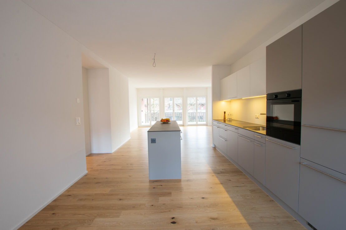 Moderne Küche mit hellen Holzböden, einer Kochinsel, grauen Schränken und eingebautem Ofen. Grosse Fenster am Ende des Raums lassen natürliches Licht in den Raum fluten. Minimalistisches Dekor in neutralen Tönen.