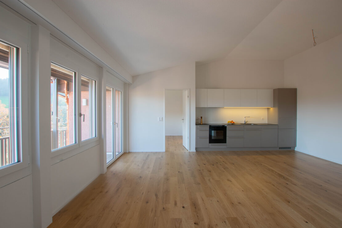 Eine geräumige, leere, moderne Küche mit hellem Holzboden und weissen Wänden. Sie verfügt über grosse Fenster auf der linken Seite, eine grauweisse Küchenzeile mit Backofen und Kochfeld und ein minimalistisches Design.