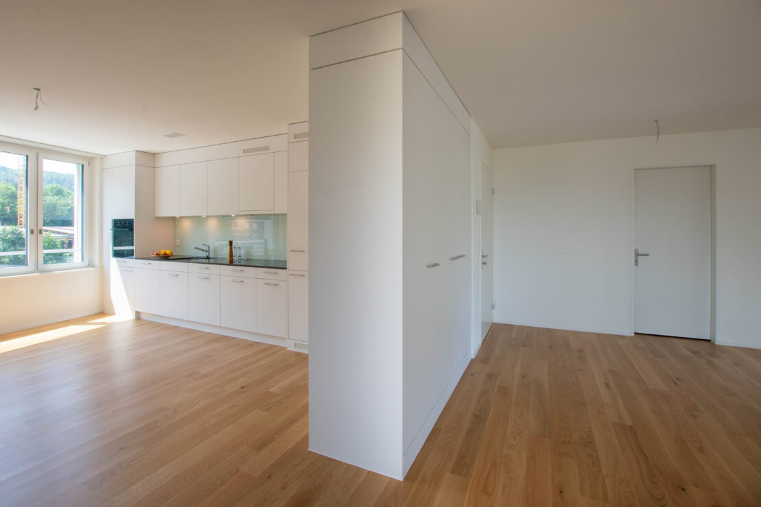 Eine moderne Küche mit weissen Schränken und hellem Holzboden. Grosse Fenster auf der linken Seite lassen natürliches Licht herein. Eine Insel trennt die Küche vom offenen Wohnbereich, der durch einen ähnlichen Holzboden und eine geschlossene weisse Tür sichtbar ist.
