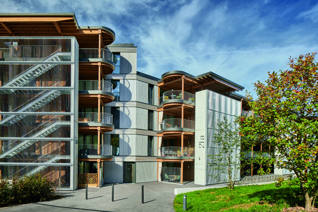Modernes Wohnhaus mit geschwungenen Balkonen und Holzfassade vor einem blauen Himmel mit dünnen Wolken. Auf der linken Seite sind Treppen zu sehen, umgeben von Grünflächen und einem gepflasterten Gehweg.