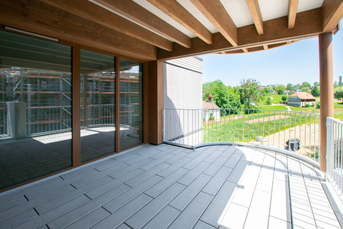 Ein geräumiger Balkon mit Holzdecke, Metallgeländer und grauem Fliesenboden. Grosse Glastüren führen hinein. Der Blick umfasst grüne Bäume, Felder und ein entferntes Haus unter einem klaren blauen Himmel.