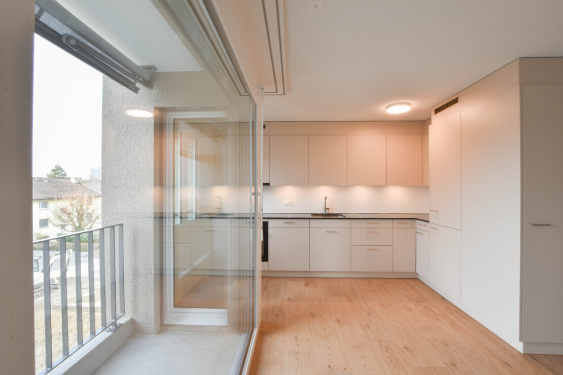 Eine moderne Küche mit beigen Schränken, schwarzen Arbeitsplatten und Einbauleuchten. Auf der linken Seite befindet sich ein grosses Fenster, das Tageslicht hereinlässt. Der Boden besteht aus hellem Holz und eine Glasschiebetür führt auf einen kleinen Balkon.
