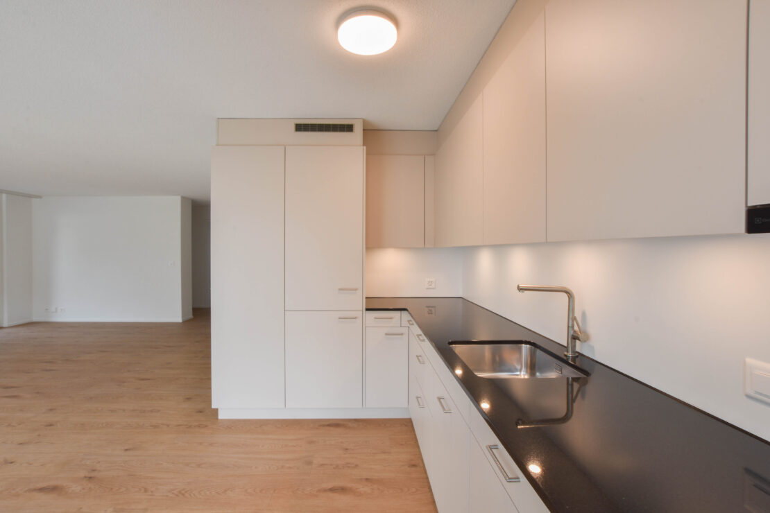 Moderne Küche mit weissen Schränken und einer eleganten schwarzen Arbeitsplatte. Spüle und Wasserhahn sind in die Arbeitsplatte integriert. Der Raum verfügt über Holzboden und wird durch eine Deckenleuchte gut beleuchtet. Im Hintergrund ist ein offener Bereich sichtbar.