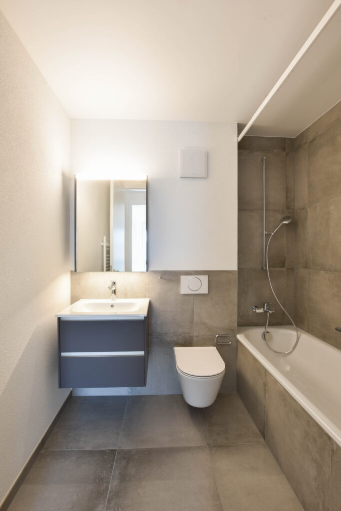 Salle de bains moderne avec des carreaux gris et un design minimaliste. Dispose d'un lavabo flottant, d'une armoire à glace, de toilettes murales et d'une baignoire avec dispositif de douche. Les murs sont clairs, ce qui renforce la sensation d'espace.