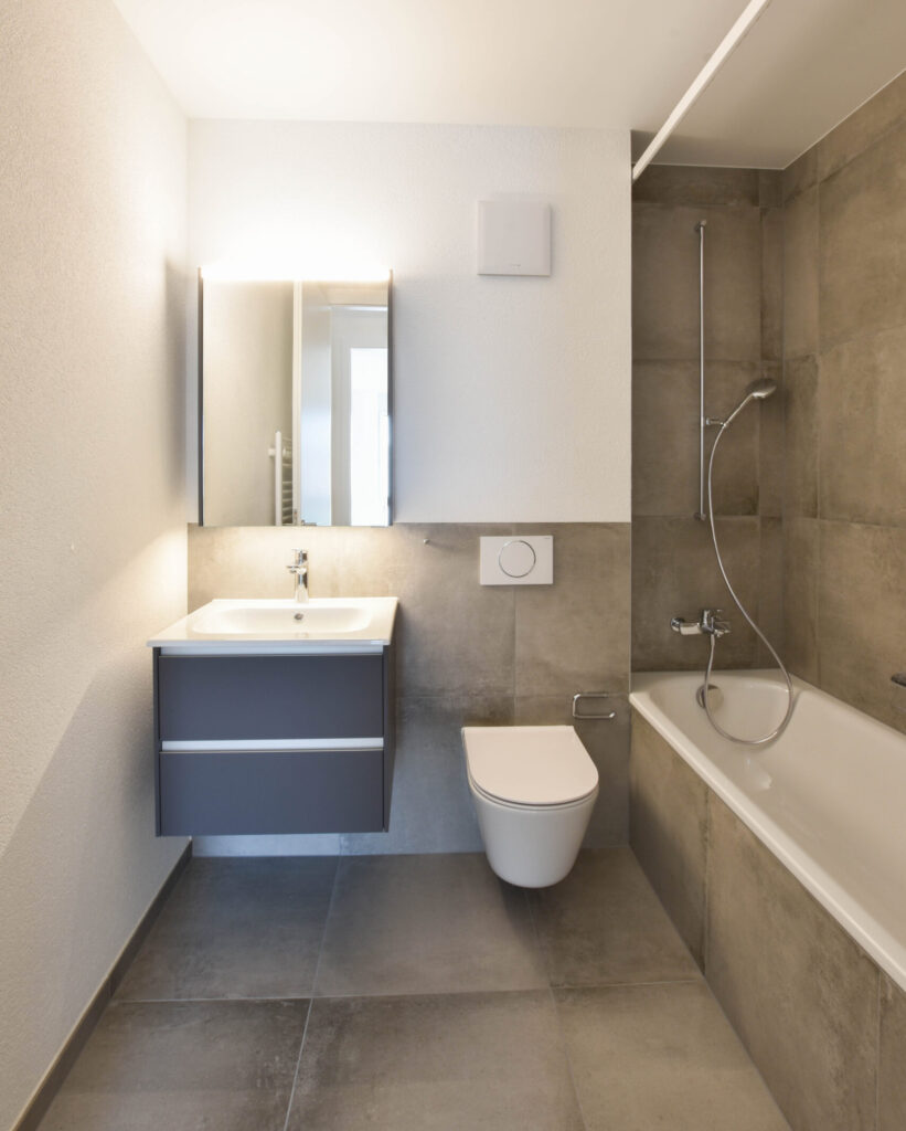 Modernes Badezimmer mit grauen Fliesen und minimalistischem Design. Verfügt über ein schwebendes Waschbecken, einen Spiegelschrank, eine wandmontierte Toilette und eine Badewanne mit Duschvorrichtung. Die Wände sind hell, was das geräumige Gefühl verstärkt.