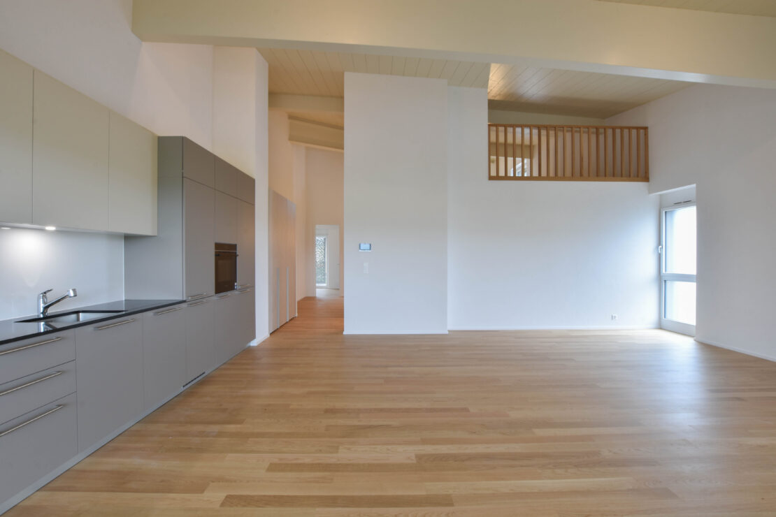 Ein geräumiger, moderner offener Raum mit hellem Holzboden, hohen Decken und Küchenschränken in neutralen Farben. Es gibt einen Balkon im Zwischengeschoss und ein grosses Fenster, das Tageslicht hereinlässt.