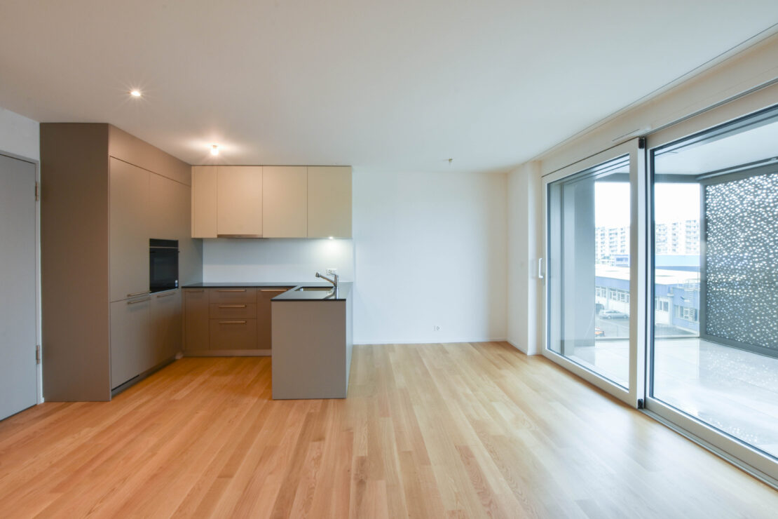 Eine moderne, unmöblierte Wohnung mit hellem Holzboden, einer kompakten Küche mit braunen und weissen Schränken und einer kleinen Kochinsel. Grosse Glastüren öffnen sich zu einem Balkon mit einem Sichtschutz aus perforiertem Metall und bieten einen Blick auf die umliegenden Gebäude.