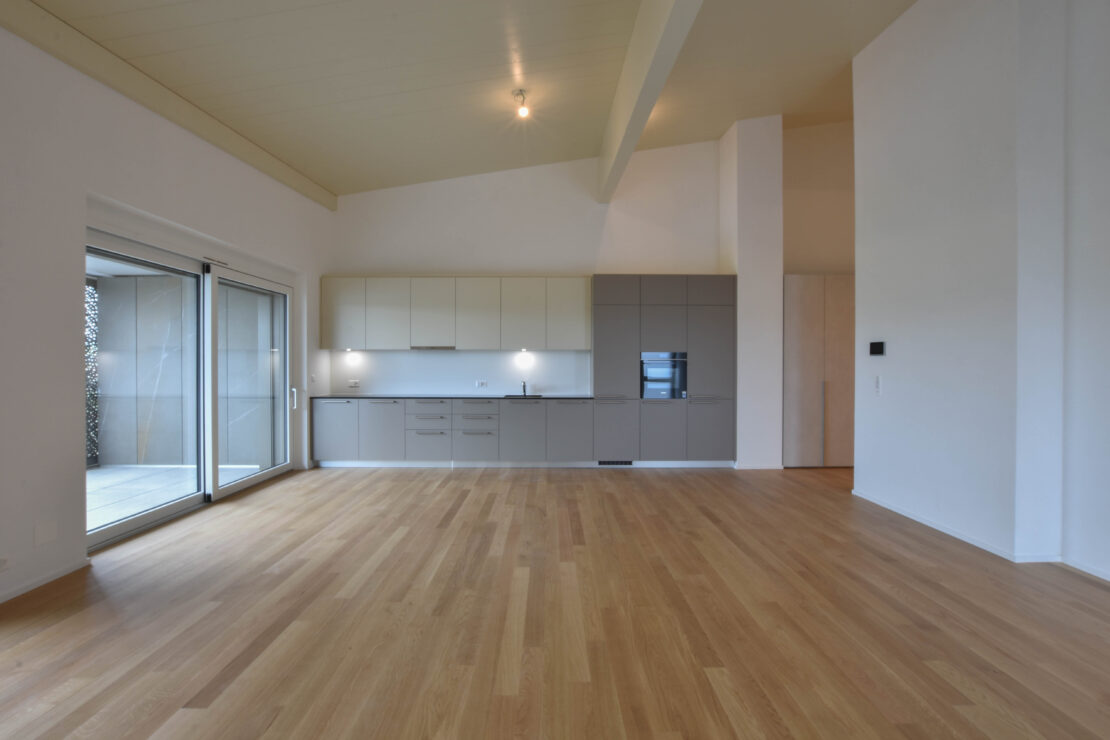 Geräumiger, unmöblierter offener Wohnbereich mit Holzboden, hohen Decken und moderner Küche mit hellen Schränken. Grosse Glasschiebetüren auf der linken Seite öffnen sich zu einem Balkon, der den Raum mit natürlichem Licht erhellt.