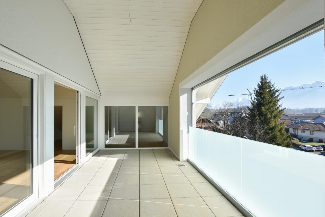 Un balcon spacieux et ensoleillé avec un sol carrelé clair, entouré de balustrades en verre. De grandes portes vitrées mènent à l'intérieur. Au loin, on aperçoit des arbres et des toits sous un ciel bleu limpide.