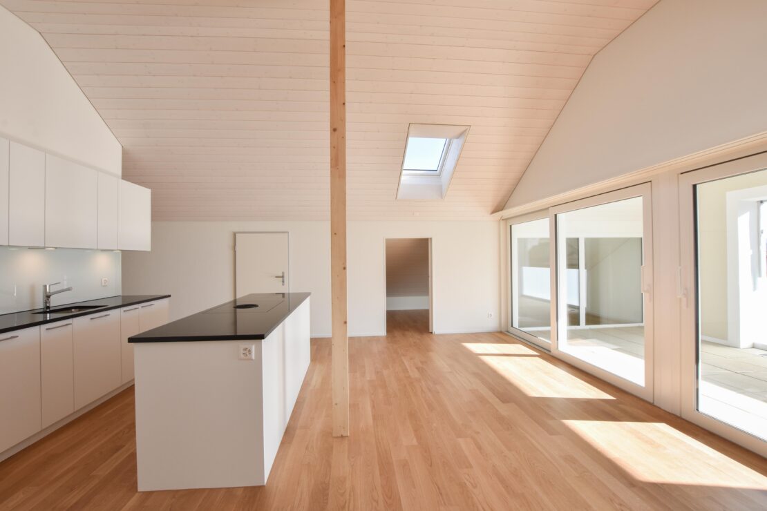 Un intérieur moderne et minimaliste avec un espace ouvert au sol en bois clair. L'espace comprend une cuisine avec des plans de travail noirs lisses et des armoires blanches, une colonne centrale en bois, de grandes portes coulissantes en verre et un puits de lumière au plafond.