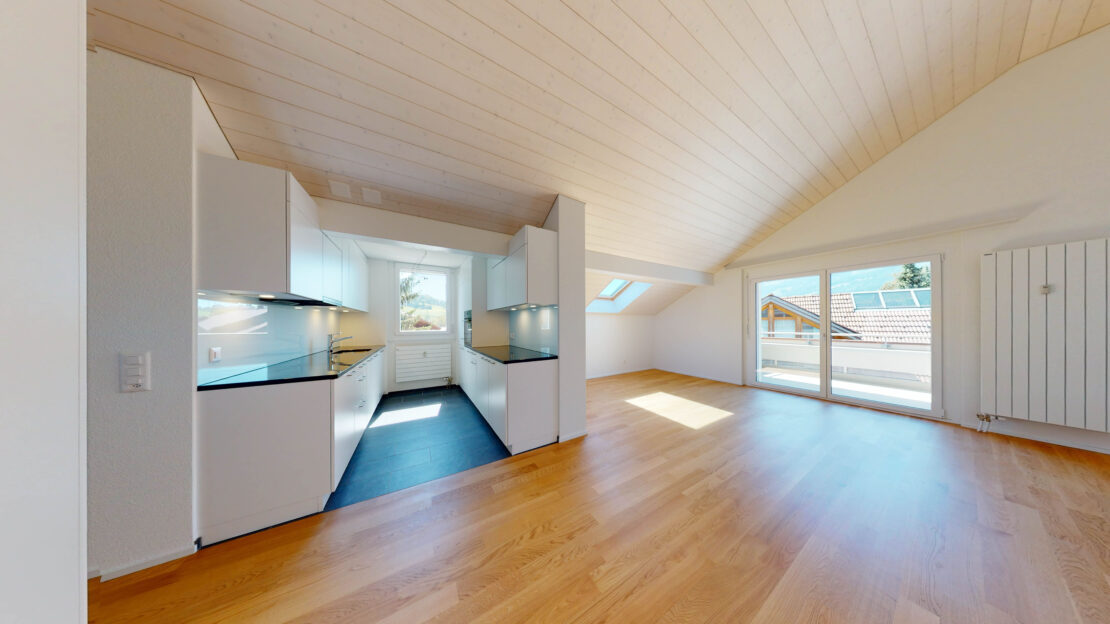 Un intérieur d'appartement lumineux et moderne dispose d'une petite cuisine avec des armoires blanches et des plans de travail noirs. La pièce a des sols en bois, un plafond incliné avec des lucarnes et de grandes fenêtres qui offrent une vue sur les toits et les environs.