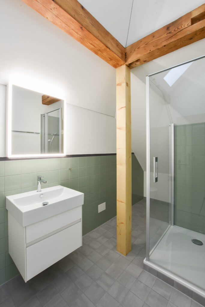 Une salle de bain moderne avec un grand miroir éclairé au-dessus d'un lavabo blanc, des murs carrelés en vert et une poutre en bois. Une cabine de douche en verre est posée sur un sol carrelé gris, au-dessus duquel se trouve une pente de toit.