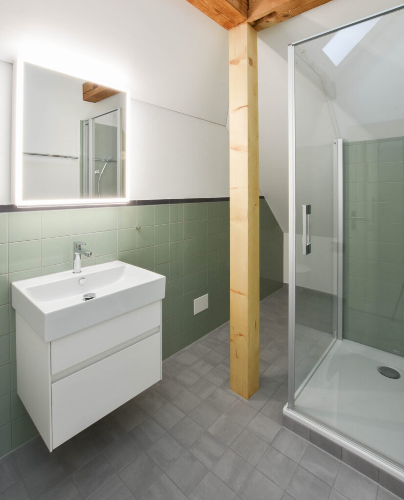 Ein modernes Badezimmer mit einem grossen beleuchteten Spiegel über einem weissen Waschbecken, grün gefliesten Wänden und einem Holzbalken. Eine Glasduschkabine steht auf einem grau gefliesten Boden, darüber befindet sich eine Dachschräge.