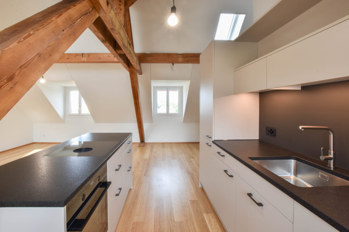 Eine moderne Küche im Dachgeschoss mit eleganten weissen Schränken, schwarzen Arbeitsplatten und Holzbalken. Sie verfügt über eine Insel mit Induktionskochfeld und Backofen, eine Spüle und grosse Fenster, die für reichlich natürliches Licht sorgen.