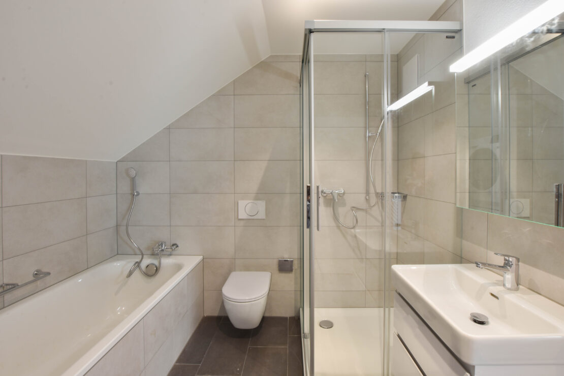 Ein modernes Badezimmer mit einer weissen Badewanne auf der linken Seite, einer wandmontierten Toilette in der Mitte und einer verglasten Dusche auf der rechten Seite. Neben der Dusche befindet sich ein elegantes weisses Waschbecken mit Spiegel. Der Raum hat hellgraue Fliesen und klare Linien.