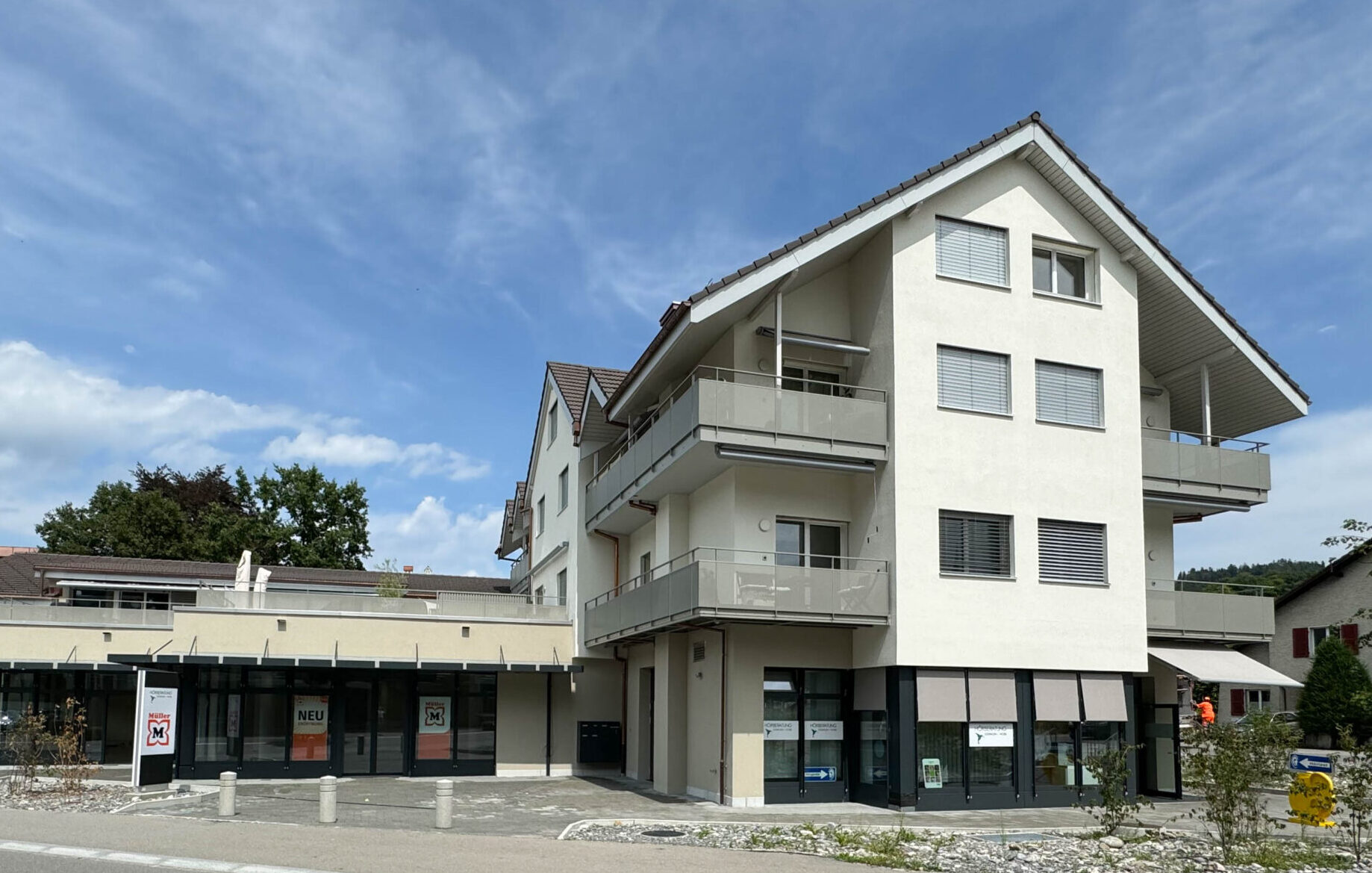 Ein modernes, mehrstöckiges Gebäude mit Balkonen und grossen Fenstern unter einem klaren blauen Himmel. Die untere Etage hat Schaufenster mit Werbung in den Fenstern. Die Umgebung umfasst eine gepflasterte Fläche und etwas Grün.