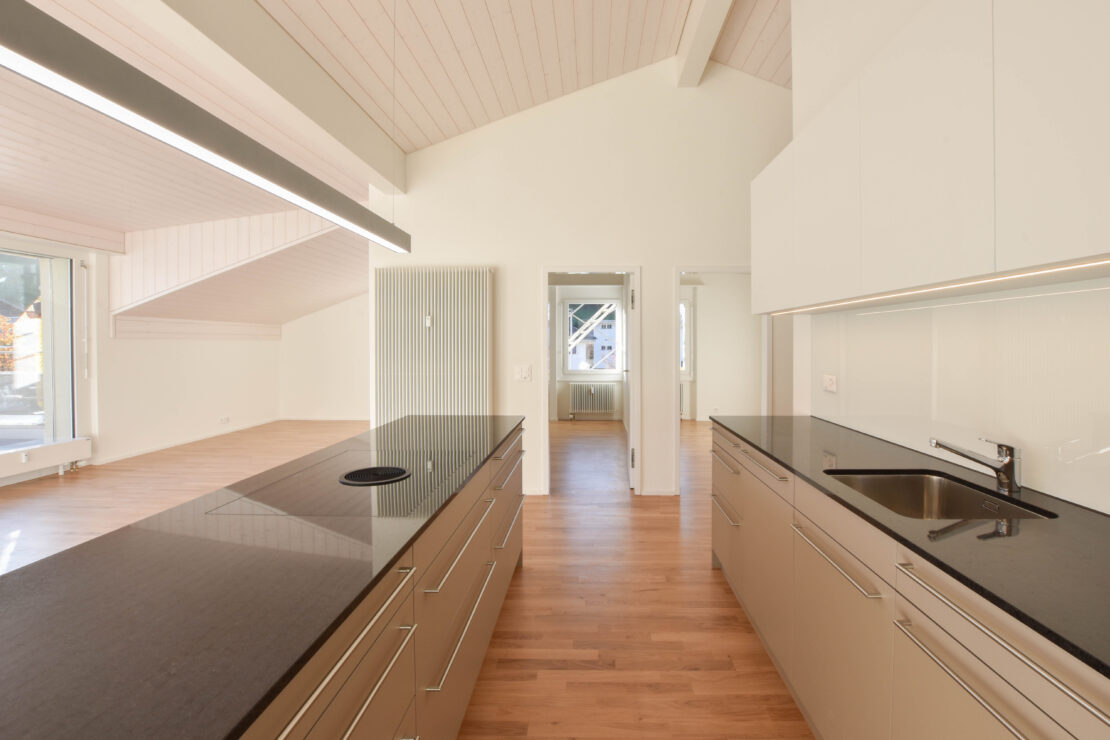 Helle, moderne Küche mit eleganten Schränken und einer glänzenden, dunklen Arbeitsplatte. Der Raum verfügt über einen hellen Holzboden, eine hohe Decke mit Holzpaneelen und viel natürliches Licht, das durch die Fenster im angrenzenden Raum fällt.