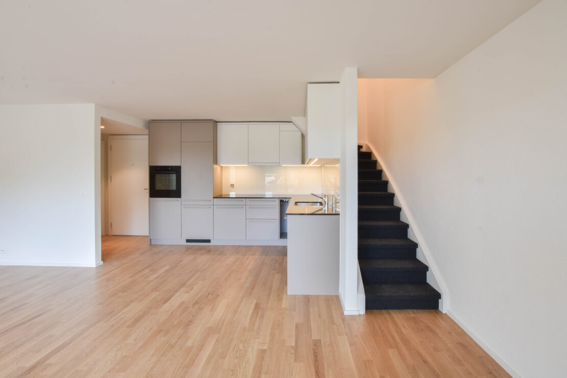 Ein geräumiges, modernes Apartment-Interieur mit hellem Holzboden. Der Raum verfügt links über eine Küche mit grauen Schränken und weissen Arbeitsplatten. Rechts führt eine schwarze Treppe nach oben. Der Raum ist mit natürlichem Licht gut beleuchtet.