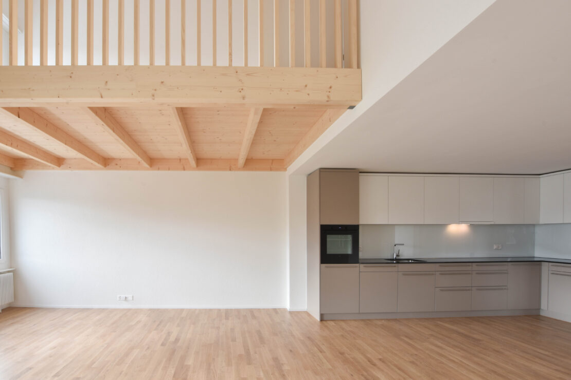 Ein geräumiger, moderner Innenraum mit hellen Holzböden und hohen Decken. Die linke Seite verfügt über grosse Fenster und einen Dachboden mit Holzgeländer darüber. Die rechte Seite hat eine minimalistische Küche mit Schränken in neutralen Tönen und integrierten Geräten.