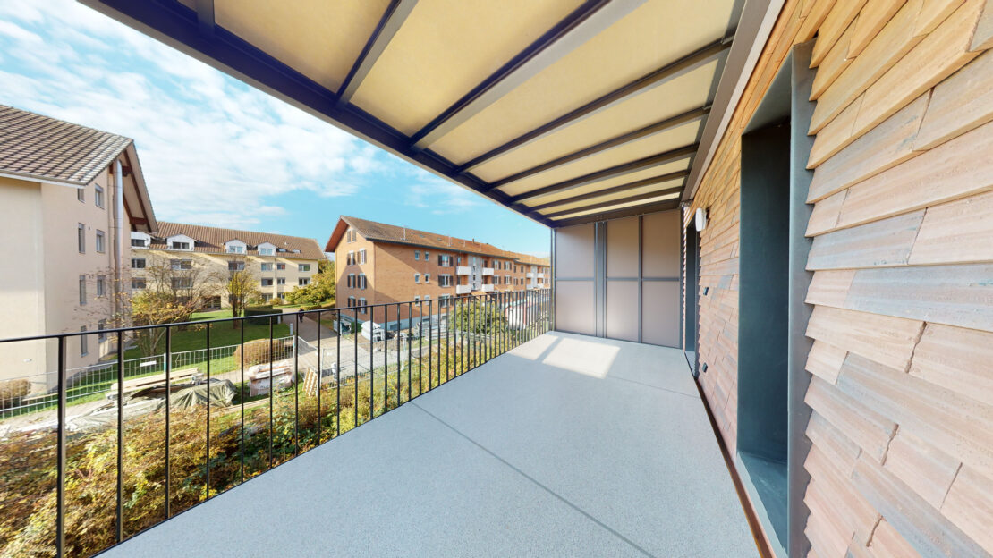 Blick von einem überdachten Balkon auf ein Vorstadtviertel mit dreistöckigen Wohnhäusern und einem Gartenbereich. Der Balkon hat ein Geländer und der Himmel ist klar mit ein paar Wolken.