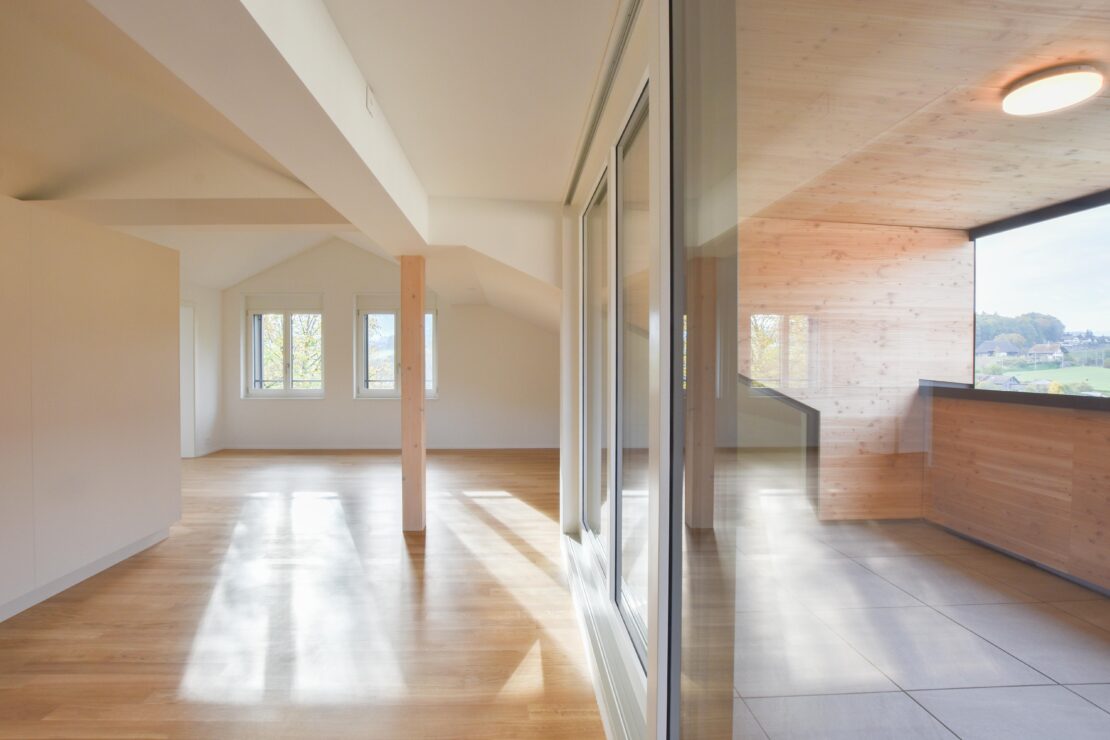 Ein modernes, geräumiges Zimmer mit grossen Fenstern, die für Tageslicht sorgen. Das Zimmer verfügt über helle Holzböden und einen Bereich mit Fliesenboden. Es gibt einen Holzbalken und die Wände sind weiss gestrichen und holzgetäfelt. Von draussen hat man eine malerische Aussicht.
