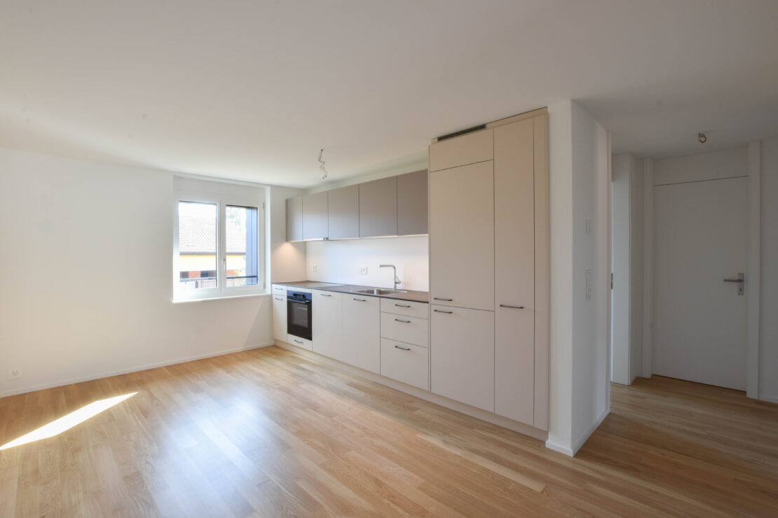 Eine moderne, leere Küche mit hellem Holzboden und beigen Schränken. Die Küche verfügt über einen integrierten Backofen und Geschirrspüler sowie ein kleines Fenster, das Tageslicht hereinlässt. Wände und Decke sind weiss und erzeugen ein sauberes und geräumiges Gefühl.