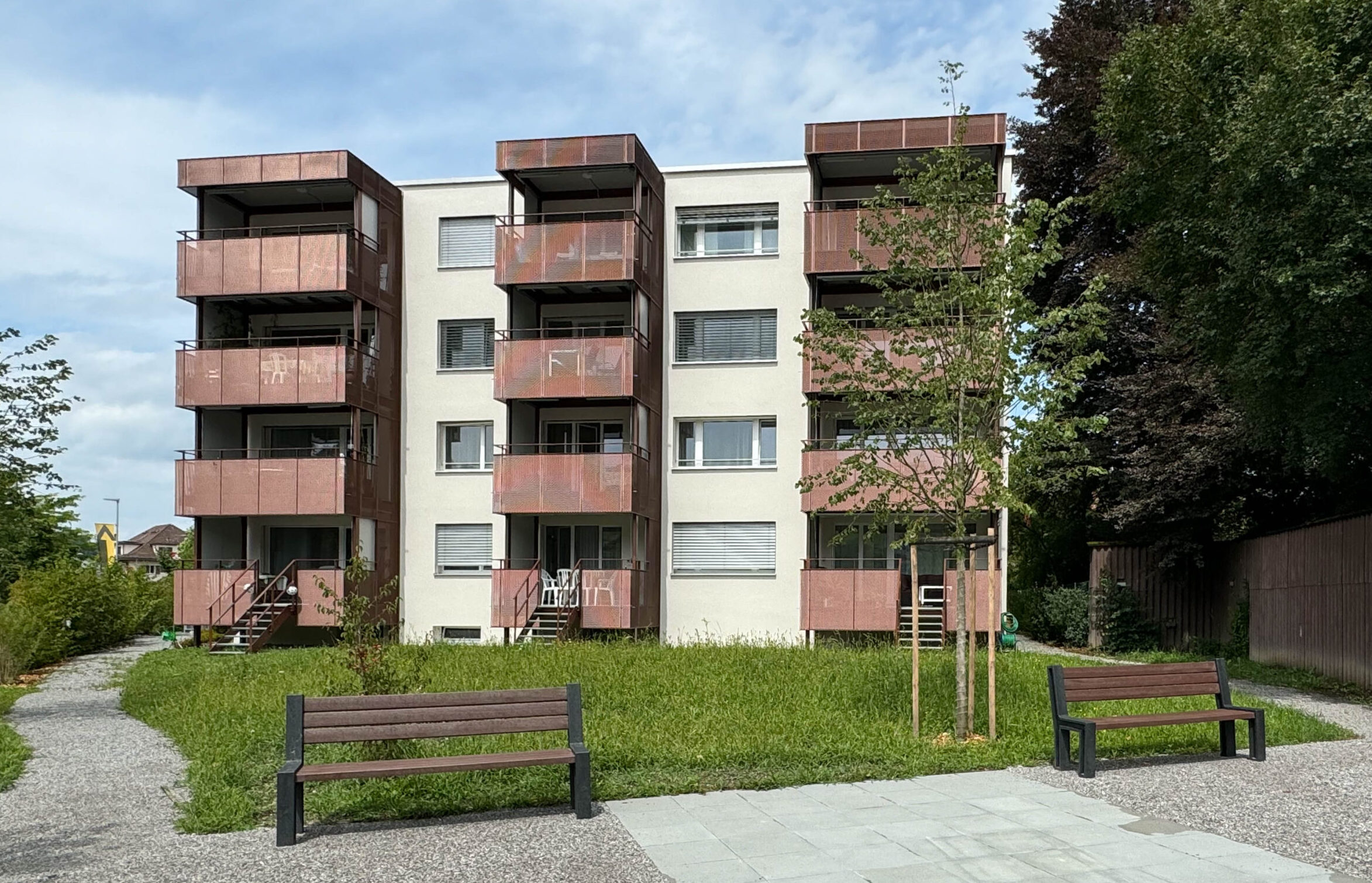 Un immeuble d'habitation de quatre étages avec des balcons bruns au milieu d'une pelouse. Deux bancs sont placés devant le bâtiment et de jeunes arbres sont plantés à proximité. Le ciel est partiellement nuageux et on peut voir des arbres verts luxuriants en arrière-plan.