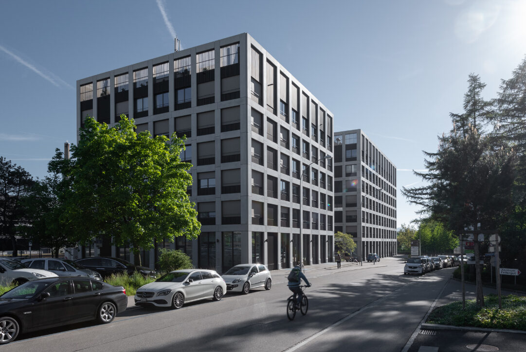 Un cycliste roule le long d'une rue ensoleillée, bordée d'immeubles de bureaux modernes et de voitures garées. Des arbres et un ciel d'un bleu éclatant remplissent l'arrière-plan et la lumière du soleil projette de longues ombres sur le trottoir.