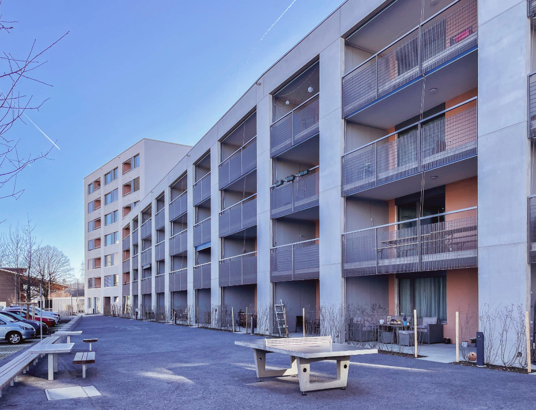 Modernes Apartmentgebäude mit mehreren Stockwerken, jedes mit Balkonen mit Geländer. Das Gebäude liegt neben einem gepflasterten Bereich mit geparkten Autos und Sitzgelegenheiten im Freien. Vereinzelte Bäume sind sichtbar und der Himmel ist klar und blau.