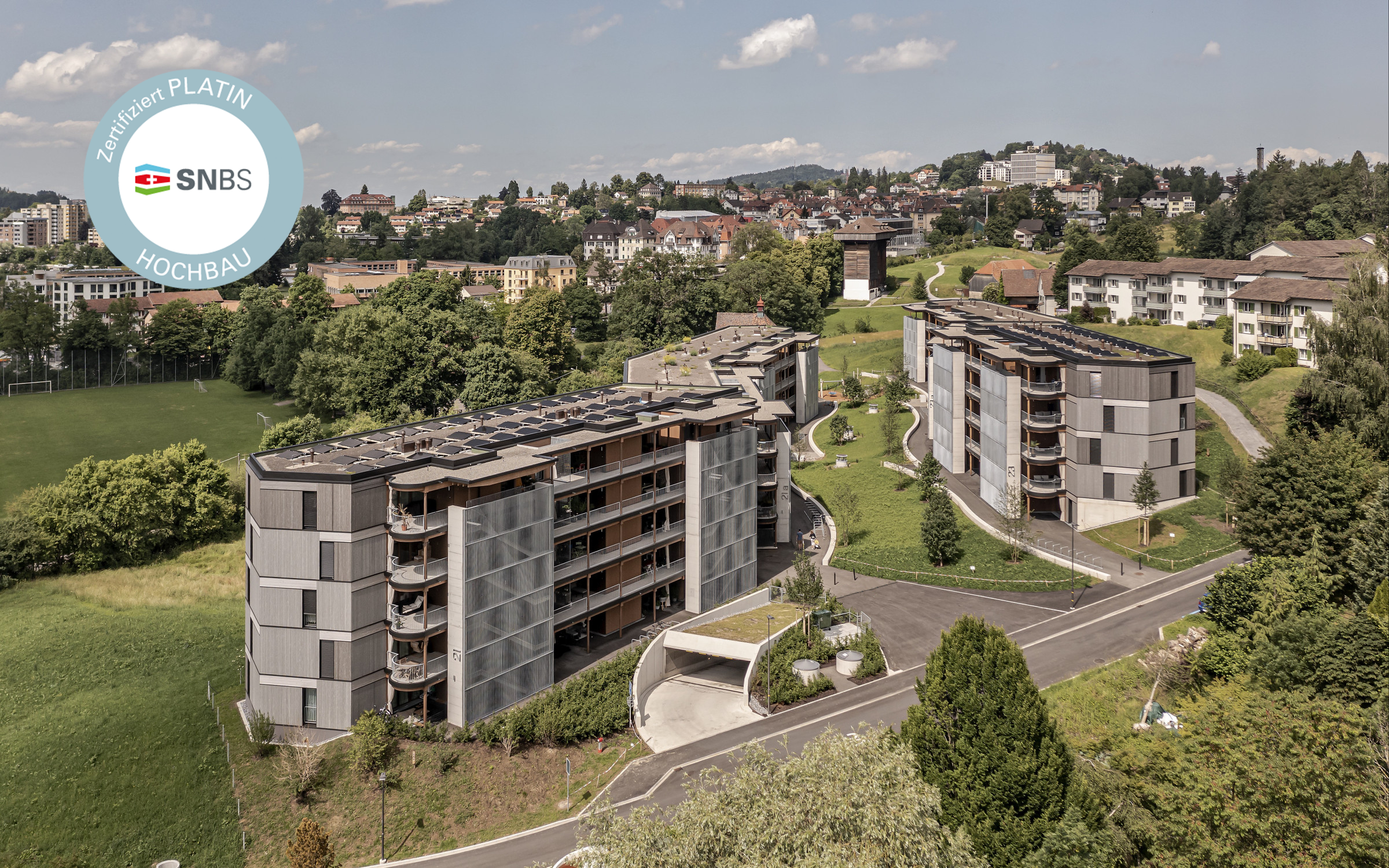 Luftaufnahme moderner Wohngebäude, umgeben von Grünflächen und einer kurvigen Strasse. Die Architektur zeichnet sich durch mehrere Balkone und Sonnenkollektoren auf den Dächern aus. In der oberen linken Ecke ist ein Zertifizierungslogo sichtbar.