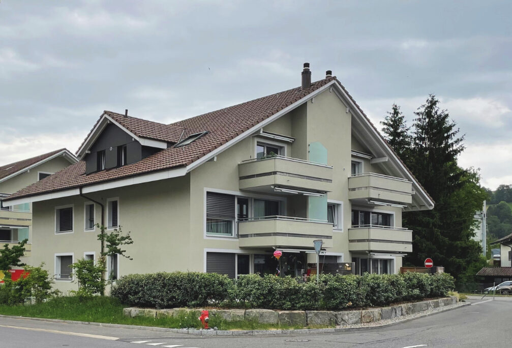 Ein modernes, mehrstöckiges Wohngebäude mit hellgrünen Wänden und braunem Dach. Es verfügt über mehrere Balkone und ist von Sträuchern umgeben. Bewölkter Himmel und Strassen mit Verkehrsschildern im Vordergrund.