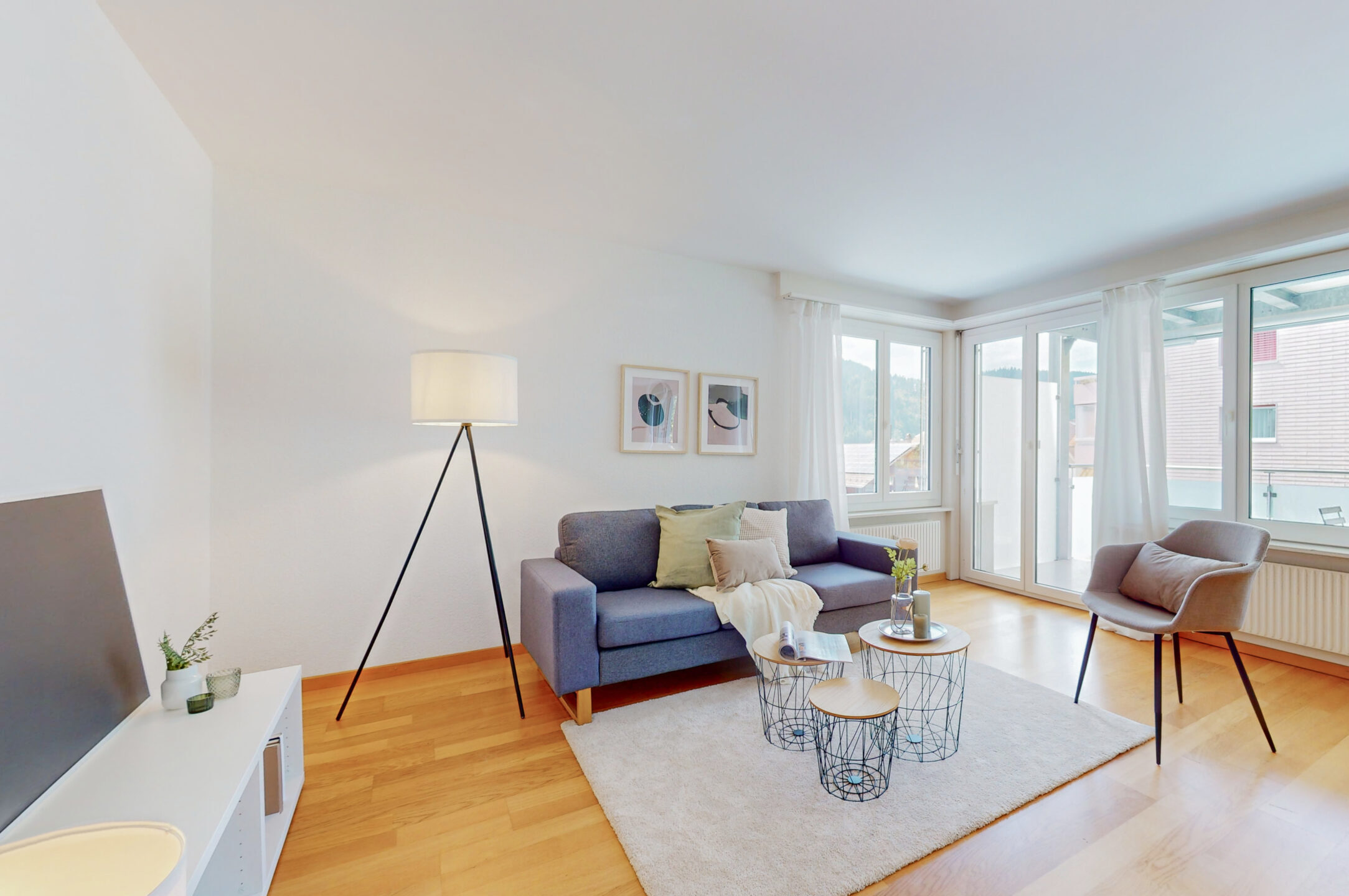 Ein modernes, gut beleuchtetes Wohnzimmer mit einem grauen Sofa, Couchtischen und einer Stehlampe. An der Wand hängen zwei gerahmte Bilder. Grosse Fenster sorgen für natürliches Licht. Ein Stuhl und minimalistisches Dekor runden den Raum ab.