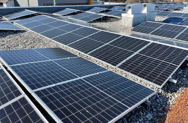 Ein Dach ist mit mehreren in ordentlichen Reihen angeordneten Solarmodulen bedeckt; die Module sind von einer Kiesfläche umgeben. Im Hintergrund sind Teile von Industrie- oder Gewerbegebäuden unter einem klaren Himmel zu sehen.